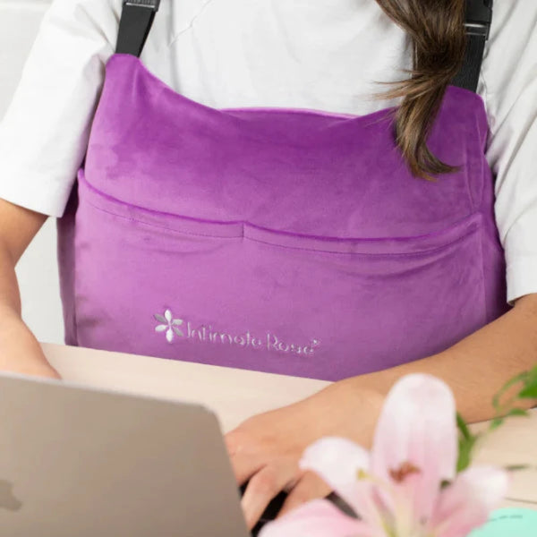 An individual is working at a laptop while wearing a vibrant purple Intimate Rose Post-Surgery Mastectomy Pillow. A blooming pink lily sits in the foreground on the table.
