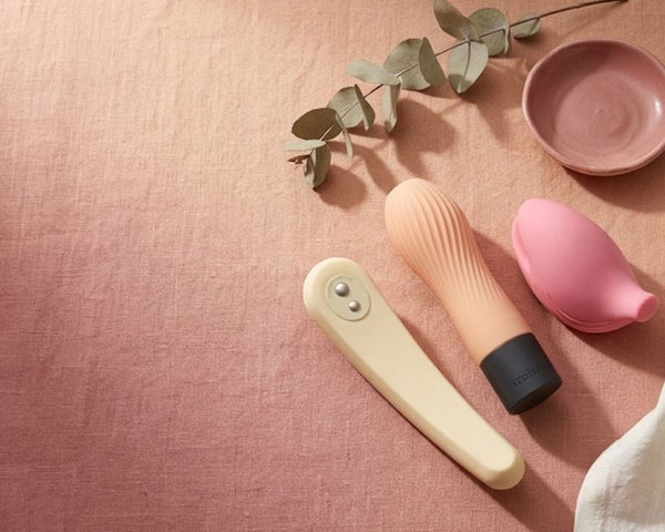 Flat-lay of three iroha soft-silicone vibrators in cream and pink on dusty-rose linen with a eucalyptus sprig