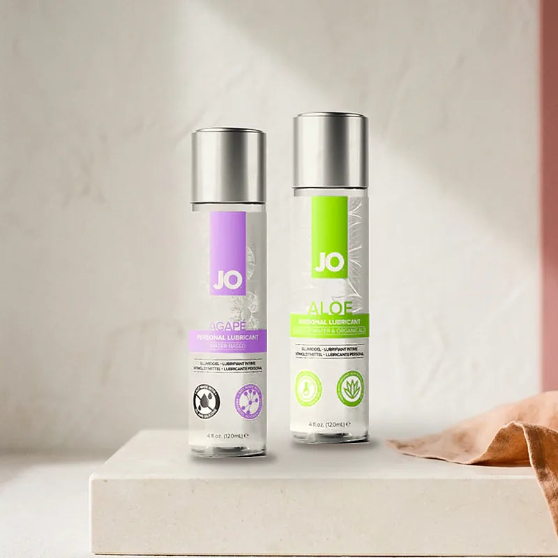 JO Agapé Original and JO Aloe water-based lubricant bottles side by side on a warm neutral surface