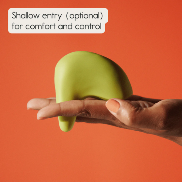 A hand holds a green, abstract, curvy object against an orange backdrop. A text bubble reads: "Shallow entry (optional) for comfort and control." It's the Kiwi Vibrating Pelvic Massager by The Pelvic People, designed for clitoral stimulation and pelvic floor relief.