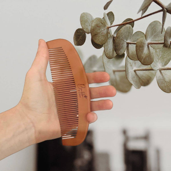 A hand holds the Bubba Bump Labour and Birth Comb by its teeth, with eucalyptus leaves subtly in view. This brown wooden comb, featuring decorative writing, provides natural pain relief and functions as an acupressure tool against a blurred, minimalistic indoor background.