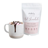 A white mug of hot chocolate with marshmallows and drizzle sits next to The Breastfeeding Tea Co’s “Lactation Hot Chocolate with Collagen” pouch—a delicious, dairy-free treat for lactation support.