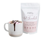 A white mug filled with hot chocolate topped with marshmallows and a chocolate drizzle sits next to a white, resealable bag labeled 