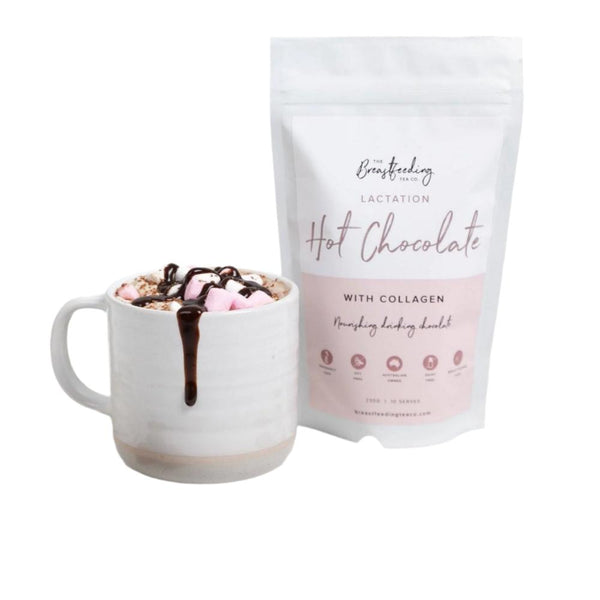 A white mug filled with hot chocolate topped with marshmallows and a chocolate drizzle sits next to a white, resealable bag labeled "Lactation Hot Chocolate with Collagen" by The Breastfeeding Tea Co. Made with organic cacao powder, the product is promoted as nourishing and supportive of breastmilk supply.