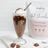 A tall glass of hot chocolate topped with whipped cream and a drizzle of chocolate, served with a silver straw. Next to the glass is a packet labeled 