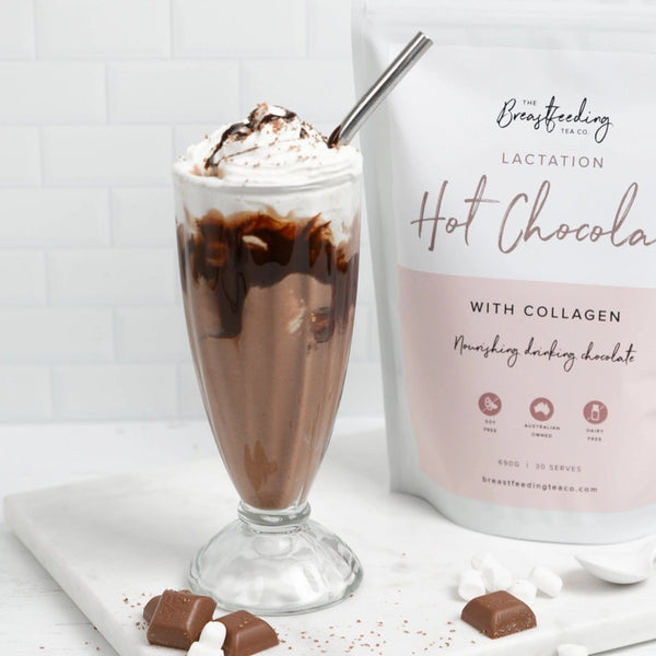 A tall glass of hot chocolate topped with whipped cream and a drizzle of chocolate, served with a silver straw. Next to the glass is a packet labeled "The Breastfeeding Tea Co Lactation Hot Chocolate with Collagen," crafted from organic cacao powder to support breastmilk supply. Scattered around are pieces of chocolate and marshmallows.