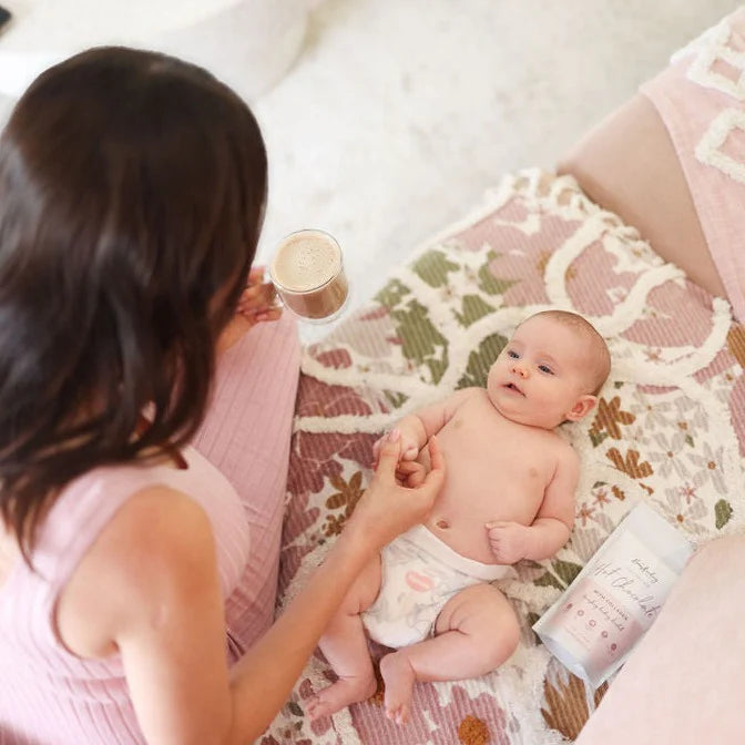 A woman in a pink dress holds a cup of coffee while sitting on a patterned blanket next to a baby wearing a white diaper. The baby lies on the blanket, looking up. Next to the baby is a package with text, possibly a snack or baby product.