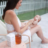 A woman in a patterned swimsuit sits outdoors on a lounge chair, smiling and holding a baby in her lap. On a small table beside her, there's a box of Lactation Tea by The Breastfeeding Tea Co with organic ingredients and a mason jar filled with tea. A white picket fence and greenery are in the background.