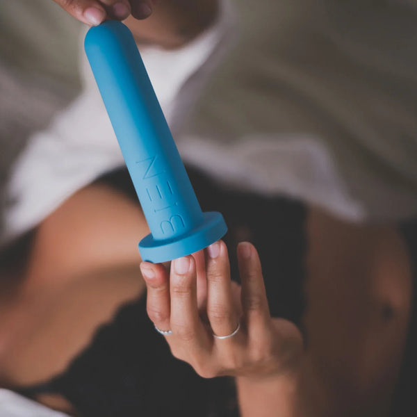 A person lying on a bed, partially covered with a white blanket, holds the Bien Silicone Vaginal Dilators - Progressive Set by BIEN Australia with both hands. They are wearing a black bra, and a thin silver ring is visible on one finger—a scene of quiet determination during post-surgery recovery.