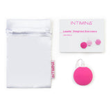 Displayed on a white background are a white drawstring pouch, user manual, and the Intimina Laselle Kegel Exercisers—round pink weighted silicone devices with loops designed to strengthen pelvic floor muscles.