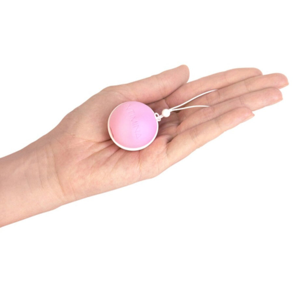 A person's outstretched hand holds an Intimina Laselle Kegel Exerciser—a small, round, pink and white weighted silicone device with an attached string loop—against a plain white background.