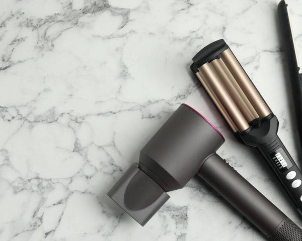 A hair styling set on a marble surface, featuring a gray hairdryer, a black and gold hair waver, and a black hair straightener.