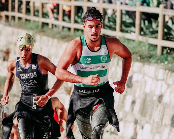 Two athletes in wetsuits and swim caps are running up a slope during a triathlon. They are focused and determined, surrounded by a stone wall and greenery in the background.