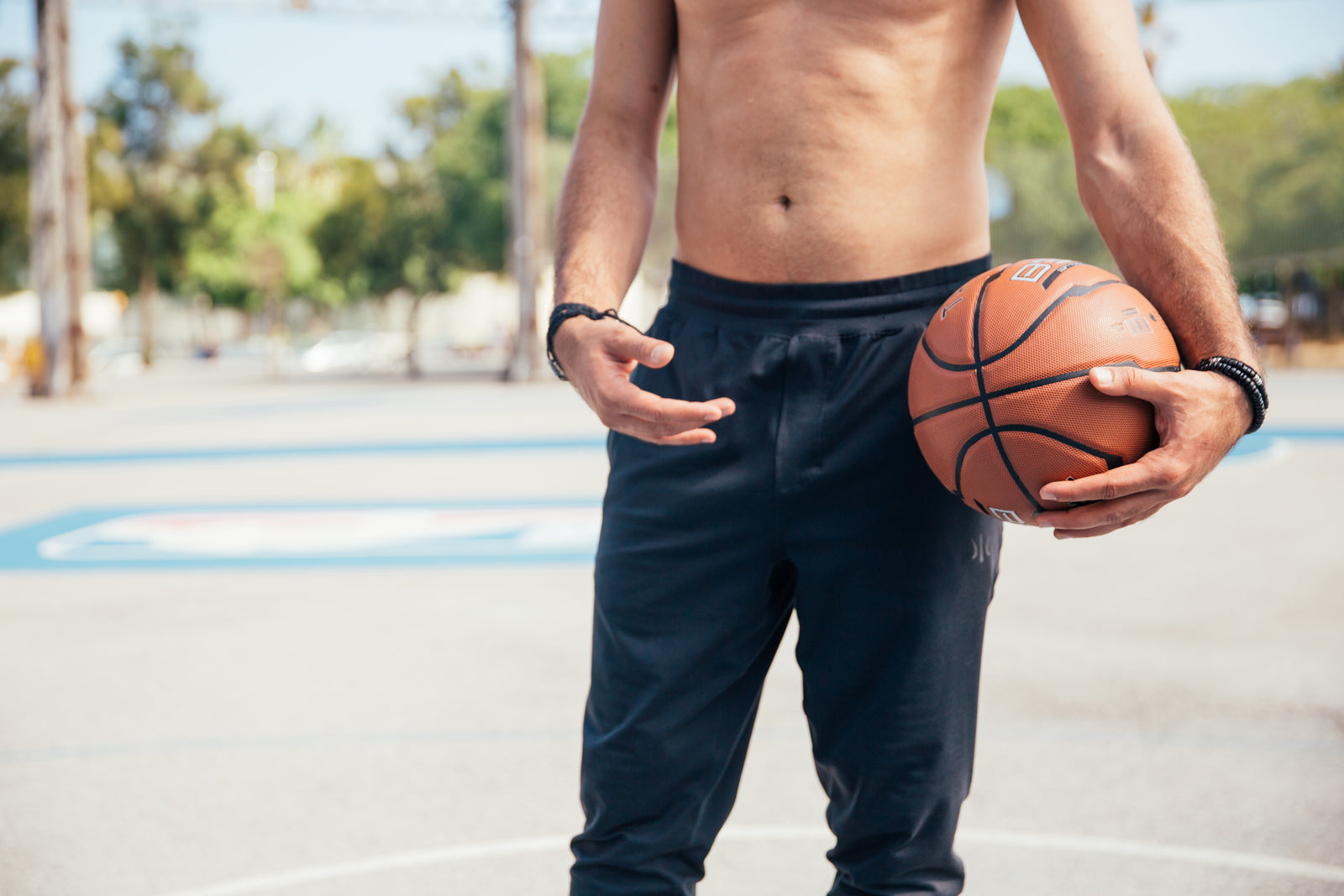 A shirtless man holding a basketball stands on an outdoor court. The background features blurred trees and light poles, creating a sunny and relaxed atmosphere.