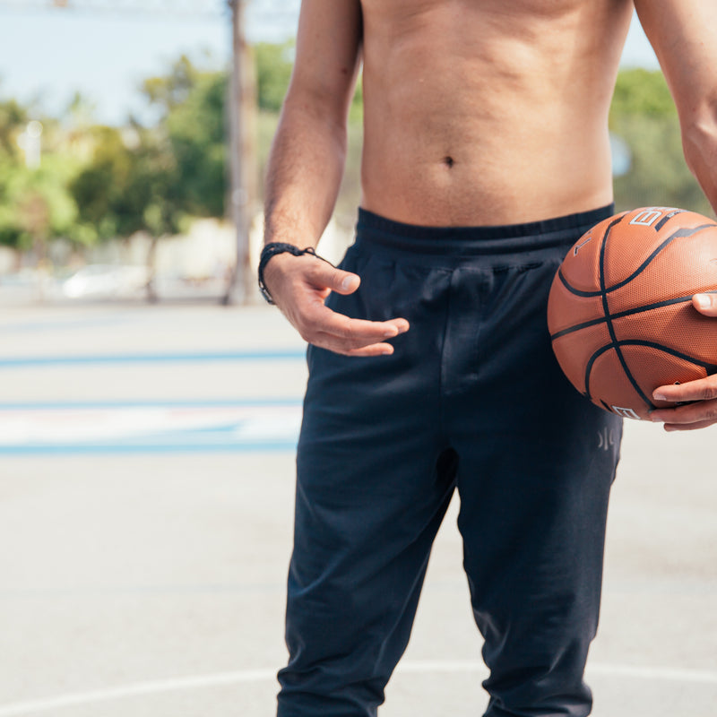 A shirtless man holding a basketball stands on an outdoor court. The background features blurred trees and light poles, creating a sunny and relaxed atmosphere.