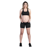 A person with long hair is standing and facing the camera, dressed in a black sports bra, SRC Maternity Shorts Mini by SRC Health, and black shoes. Their hands rest on their hips in a relaxed stance against a plain white background.