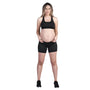 A person with long hair is standing and facing the camera, dressed in a black sports bra, SRC Maternity Shorts Mini by SRC Health, and black shoes. Their hands rest on their hips in a relaxed stance against a plain white background.