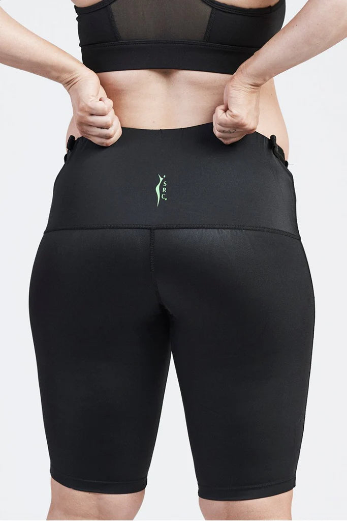 A person is shown from the back, wearing black, high-waisted SRC Maternity Shorts and a matching sports bra. They are pulling up the sides of the shorts with both hands. There is a small green SRC Health logo on the back of these maternity compression shorts designed for pelvic support during pregnancy.