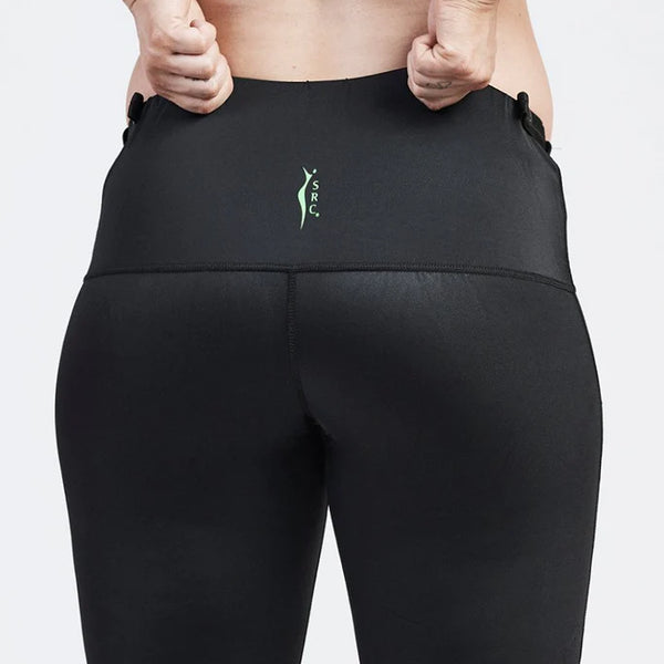 A person is shown from the back, wearing black, high-waisted SRC Maternity Shorts and a matching sports bra. They are pulling up the sides of the shorts with both hands. There is a small green SRC Health logo on the back of these maternity compression shorts designed for pelvic support during pregnancy.