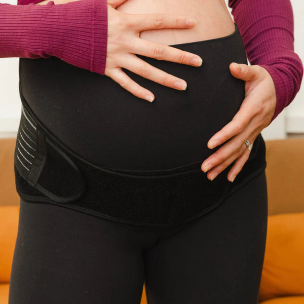 A pregnant person in a burgundy top wears the GRDL Pregnancy Belt by GRDL, sitting on a brown couch and gently cradling their belly, enjoying trusted pelvic and back support for pregnancy.