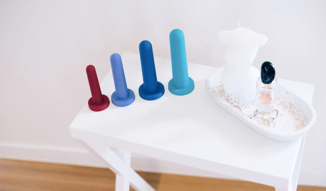 The Bien Silicone Vaginal Dilators - Medium Set from BIEN Australia, featuring an array of four vibrant dilators crafted from medical-grade silicone and available in graduated sizes, is elegantly arranged on a white table next to a decorative tray holding a torso-shaped candle, a small bottle, and rings. This setup is placed on a wooden floor against a white wall.