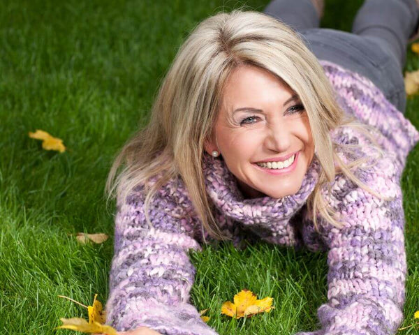 A woman with blonde hair is lying on her stomach on a grassy field, wearing a purple sweater. She is smiling and holding yellow leaves in her hands, surrounded by scattered autumn leaves.