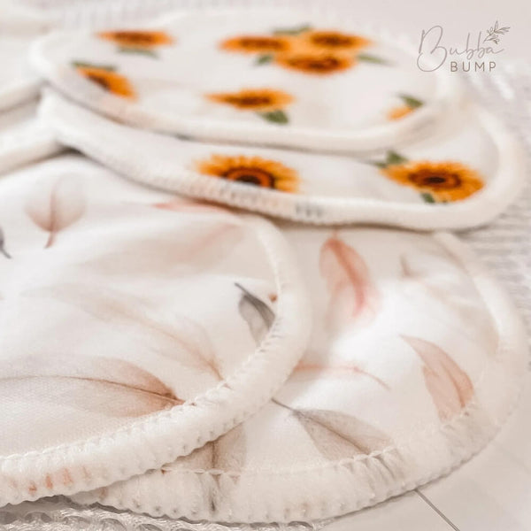 A close-up image of several soft, rounded fabric pads in various floral prints. The Organic Bamboo Reusable Breast Pads - 14 Pack are ultra-absorbent, featuring sunflowers and delicate, pale leaves. A partial logo and text "Bubba Bump" are visible in the upper right-hand corner.