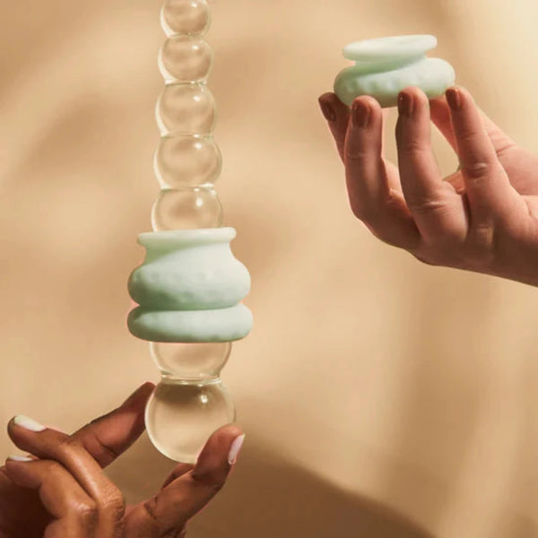 Two hands hold translucent glass objects with mint green, ribbed silicone sections. One hand cradles a large object featuring stacked spheres that resemble The Pelvic People's Ohnut Depth-Limiting Rings, while the other holds a smaller piece. The background is a neutral beige, suggesting customizable solutions for pain-free intimacy.