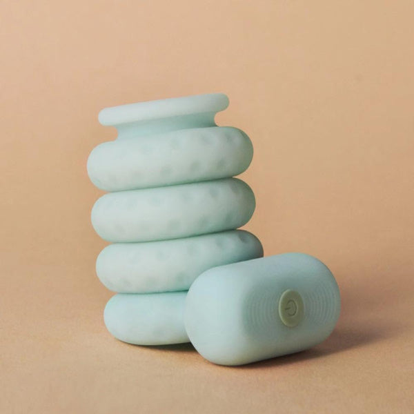 Displayed against a neutral beige background is the Ohnut Vibrating Ring from The Pelvic People, featuring a textured, light teal massage roller accompanied by a capsule-shaped device with a power button on top, intended for enhanced pleasure and pain relief.