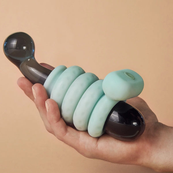A hand holding a black and teal sculptural object with a curvy form, resembling an Ohnut Vibrating Ring by The Pelvic People, features rounded shapes coiled around it, set against a neutral background.