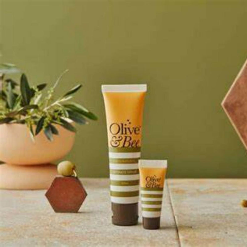 Two tubes of YES® Olive & Bee Intimate Cream—celebrated for its 100% natural ingredients—are displayed on a textured surface in front of an olive branch. The larger and smaller tubes both feature yellow and white striped packaging. In the background, a potted olive plant sits against a green backdrop.