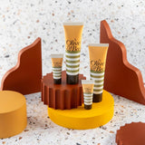 Four tubes of YES® Olive & Bee Intimate Cream are showcased. Two larger tubes and two smaller tubes, featuring 100% natural intimate cream, are placed on a yellow platform with brown, yellow, and terracotta wavy decorations in the background, set against a terrazzo-patterned surface.