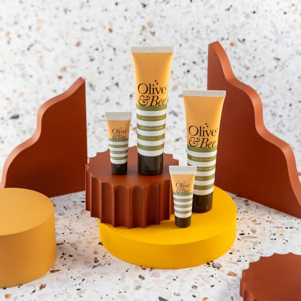 Four tubes of YES® Olive & Bee Intimate Cream are showcased. Two larger tubes and two smaller tubes, featuring 100% natural intimate cream, are placed on a yellow platform with brown, yellow, and terracotta wavy decorations in the background, set against a terrazzo-patterned surface.