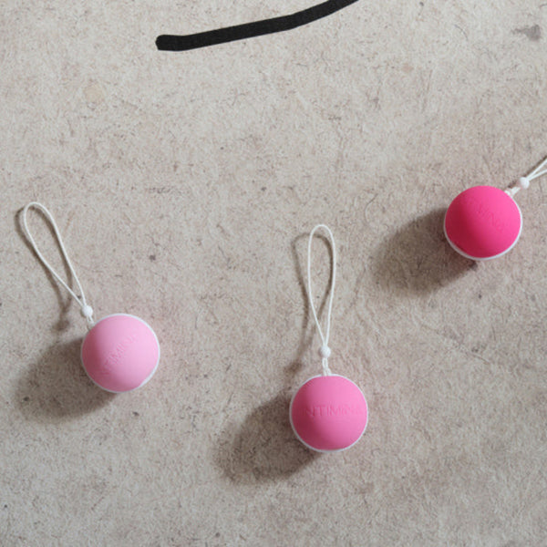 Three small, round, pink Laselle Kegel Exercisers by Intimina, each with a white string attached, are spaced apart on a textured beige surface.