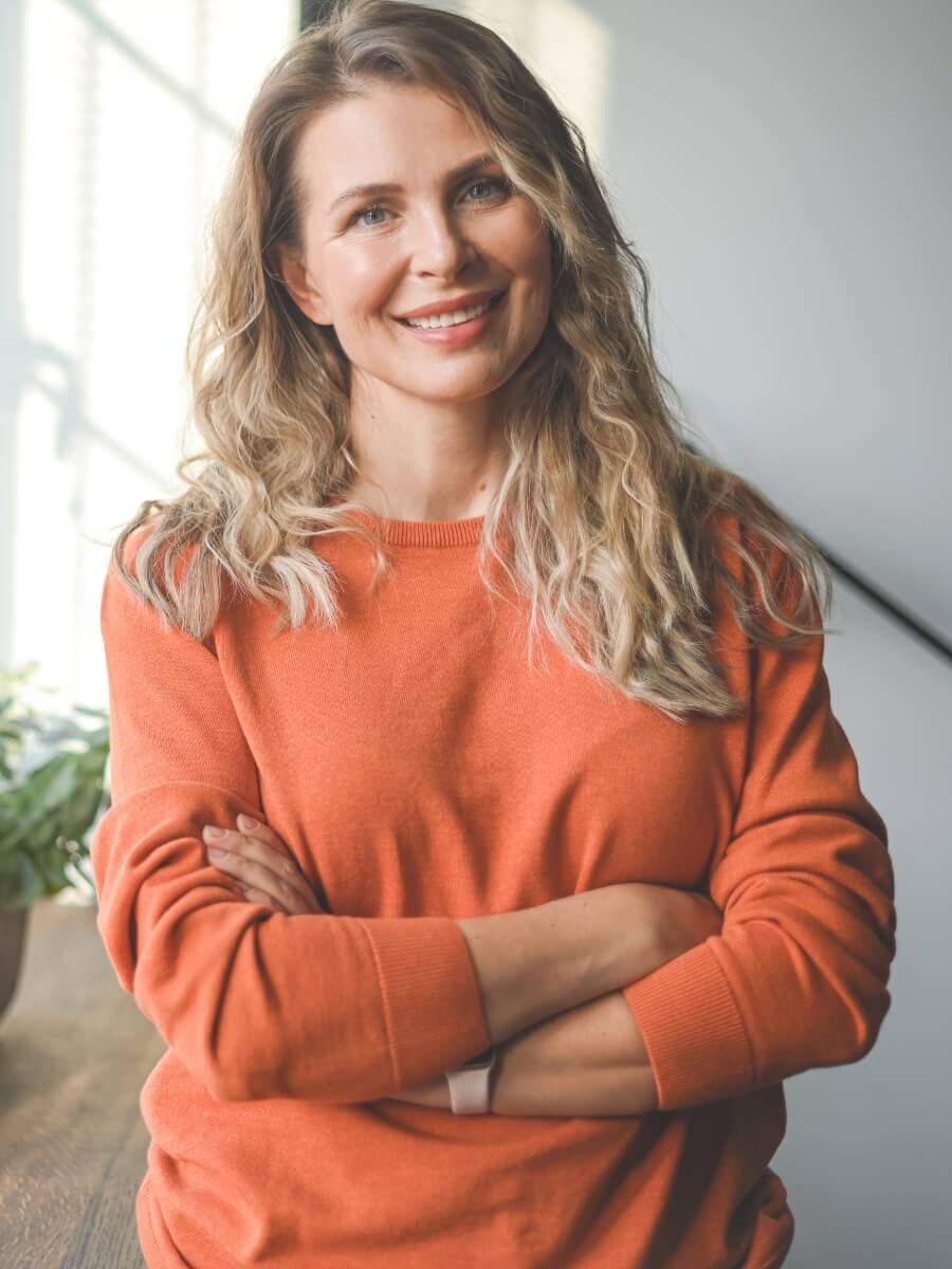 A person with long, wavy blonde hair is smiling and standing with their arms crossed. They are wearing an orange sweater. The background appears to be indoors with soft natural light and a plant visible in the corner.