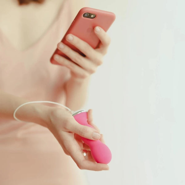 A person wearing a light pink top holds a smartphone in one hand and Perifit's ergonomically designed Perifit Care Kegel exerciser in the other. The soft, out-of-focus background complements the scene, highlighting their dedication to pelvic floor strengthening.