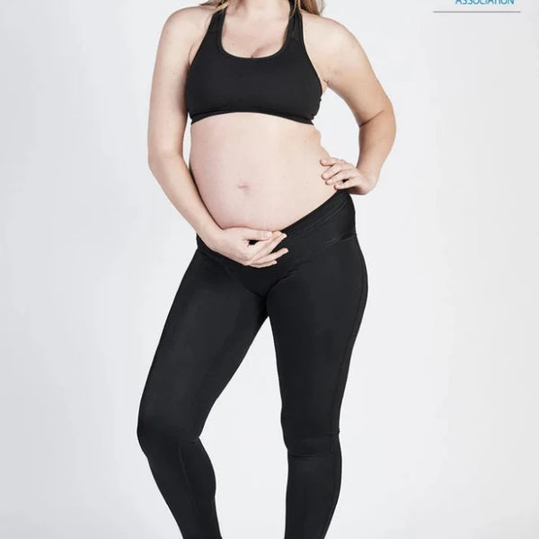 A pregnant woman is standing and smiling at the camera, dressed in a black sports bra, SRC Maternity Leggings by SRC Health in black, and black sneakers. Her left hand supports her belly while her right hand rests on her hip. The image is endorsed by the Australian Physiotherapy Association.