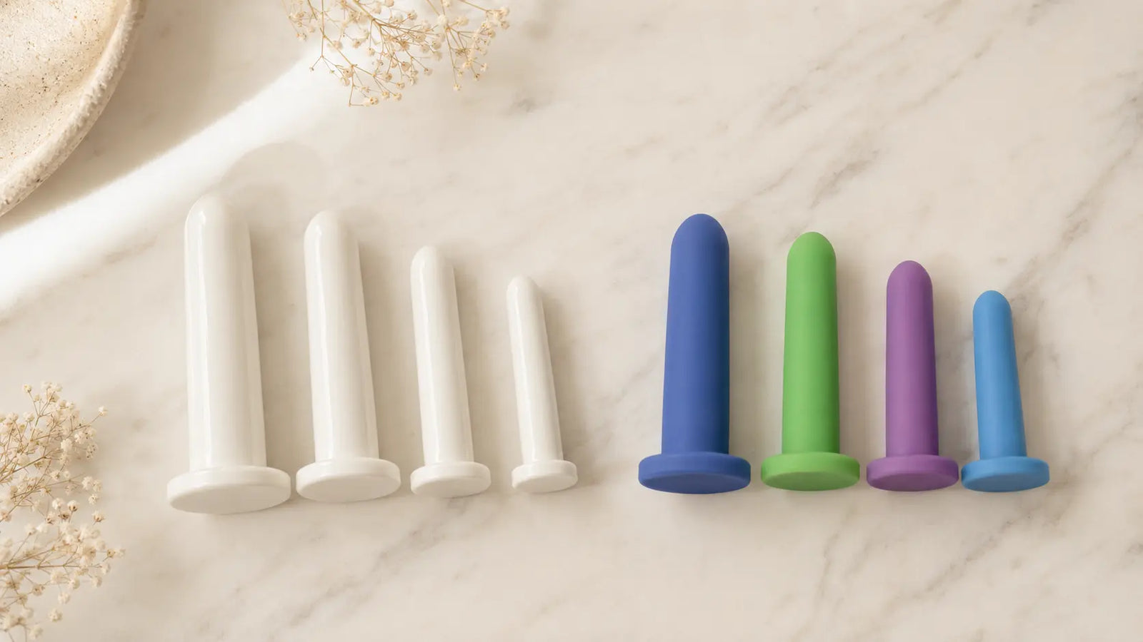 Plastic vs silicone vaginal dilators compared side by side — rigid white plastic dilators on the left and soft pink medical-grade silicone dilators on the right, on cream linen. Editorial flat-lay for pelvic floor therapy material guide.