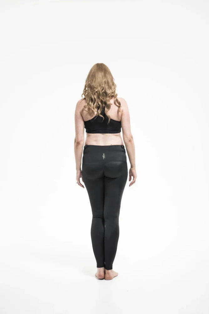 A person with long, wavy blonde hair wearing SRC Health's SRC Maternity Leggings and a black sports bra stands facing away from the camera on a plain white background.
