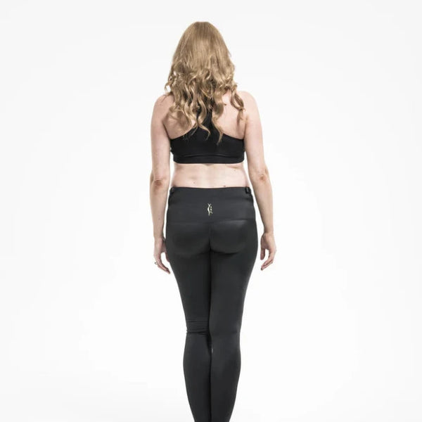 A person with long, wavy blonde hair wearing SRC Health's SRC Maternity Leggings and a black sports bra stands facing away from the camera on a plain white background.