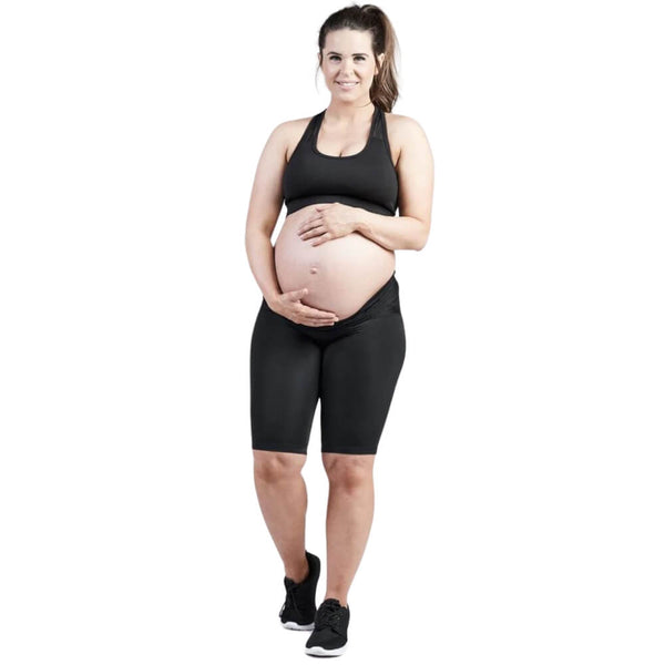 A pregnant woman wearing an SRC Health black sports bra and SRC Maternity Shorts stands with one hand on her belly and the other resting on her hip. She has long hair tied back in a ponytail and is smiling. She is also wearing black athletic shoes, against a white background.