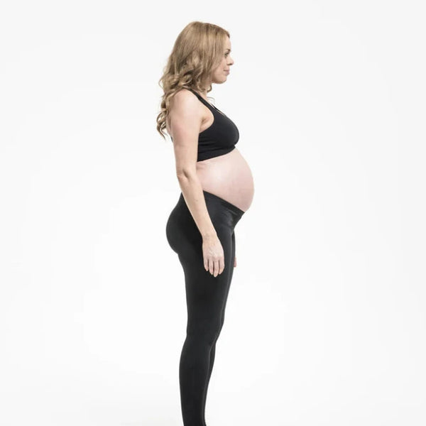 A pregnant person stands in a side profile against a plain, white background. They are wearing SRC maternity Leggings by SRC Health and a black sports bra, with bare feet, and their hair falls loosely over their shoulders.