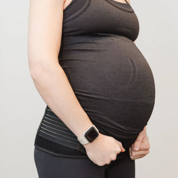 A pregnant person wearing a dark tank top, pants, and a smartwatch stands sideways, holding the GRDL Pregnancy Belt for trusted pelvic and back support by GRDL near their waist to support their baby bump.