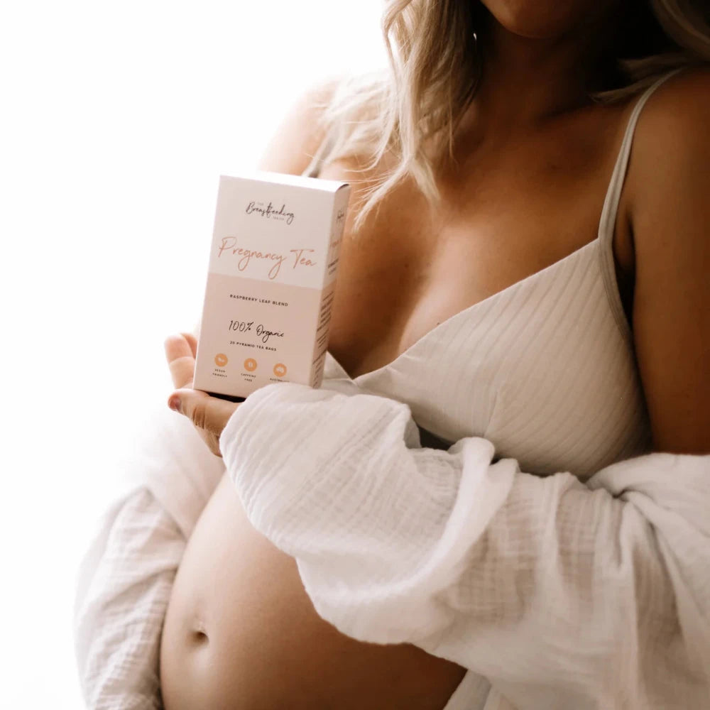 A person with blonde hair wearing a white top and a light robe holds a box of "Pregnancy Tea - Raspberry Leaf Tea" from The Breastfeeding Tea Co, featuring pregnancy-safe herbs and raspberry leaf tea, positioned in front of their bare baby bump.