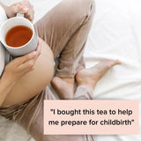A pregnant person sits on a bed, holding a mug of The Breastfeeding Tea Co's Pregnancy Tea - Raspberry Leaf Tea. They are wearing comfortable beige pants and a white top. A text box in the image reads, 
