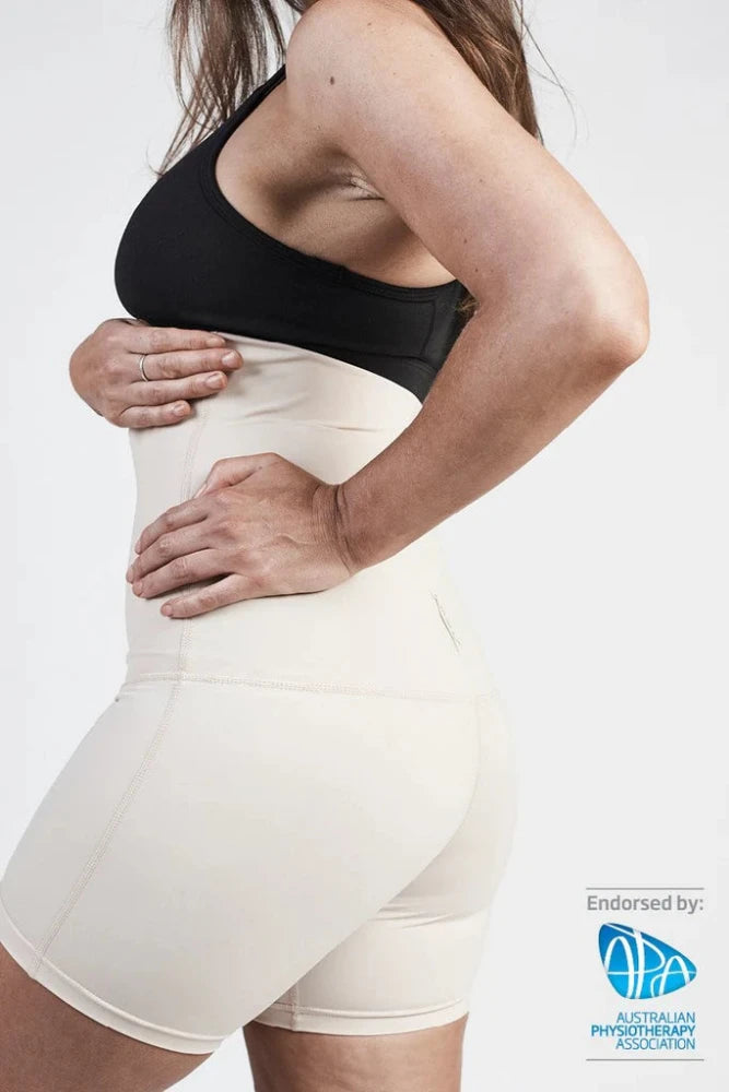 A woman wearing a black sports bra and the SRC Recovery Short - Mini in beige, from SRC Health, is shown in a side profile view with her hand placed on her back to emphasize postpartum recovery. The image is endorsed by the Australian Physiotherapy Association, as indicated by their logo in the lower right corner.