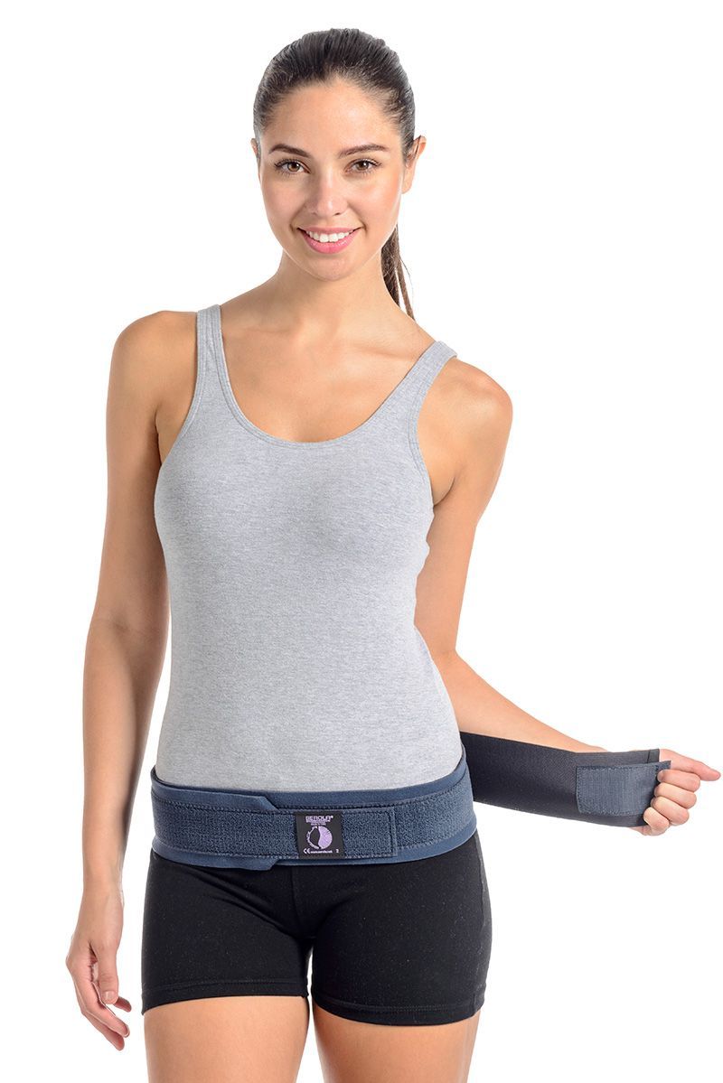 A woman wearing a gray tank top and black shorts stands smiling while fastening a black back support brace around her waist with a Velcro strap. Her hair is tied back. The background is plain white.