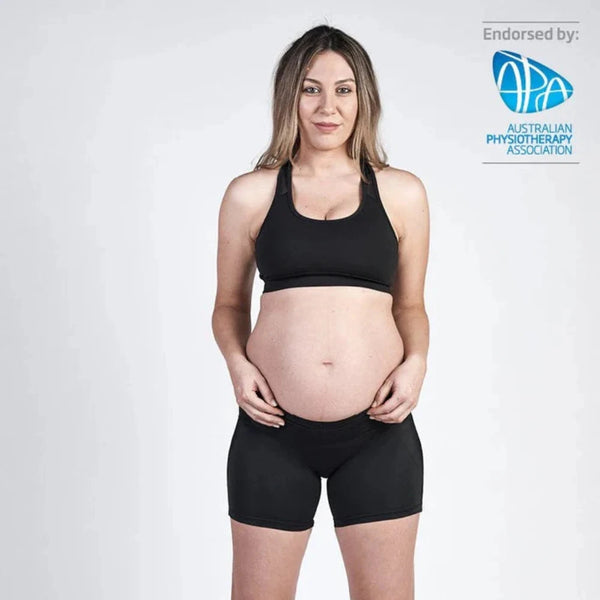 A woman stands facing forward wearing a black sports bra and SRC Maternity Shorts Mini from SRC Health, proudly showcasing her pregnant belly. She has long hair and is wearing black sneakers. The image, endorsed by the Australian Physiotherapy Association as indicated by a logo in the top right corner, highlights pregnancy support.