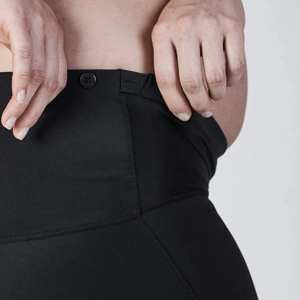 Close-up of a pregnant person adjusting the waistband of black SRC Maternity Shorts Mini by SRC Health with their hands, designed for pregnancy support. A small part of the top and belly is visible. The shorts, endorsed by the Australian Physiotherapy Association, help alleviate pelvic girdle pain, as indicated by a logo in the top right corner.
