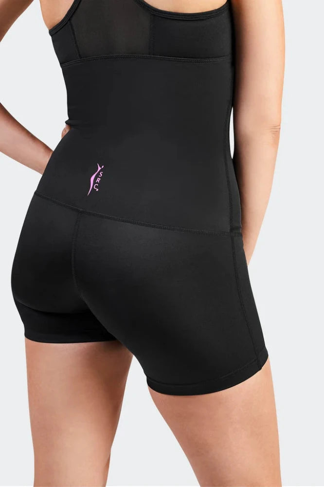 A person wearing the SRC Health's fitted black athletic outfit, specifically the SRC Recovery Short - Mini, designed for medical-grade compression. The shorts feature a small purple logo on the left side. With their left hand resting on their hip and facing away, they stand against a light gray background.